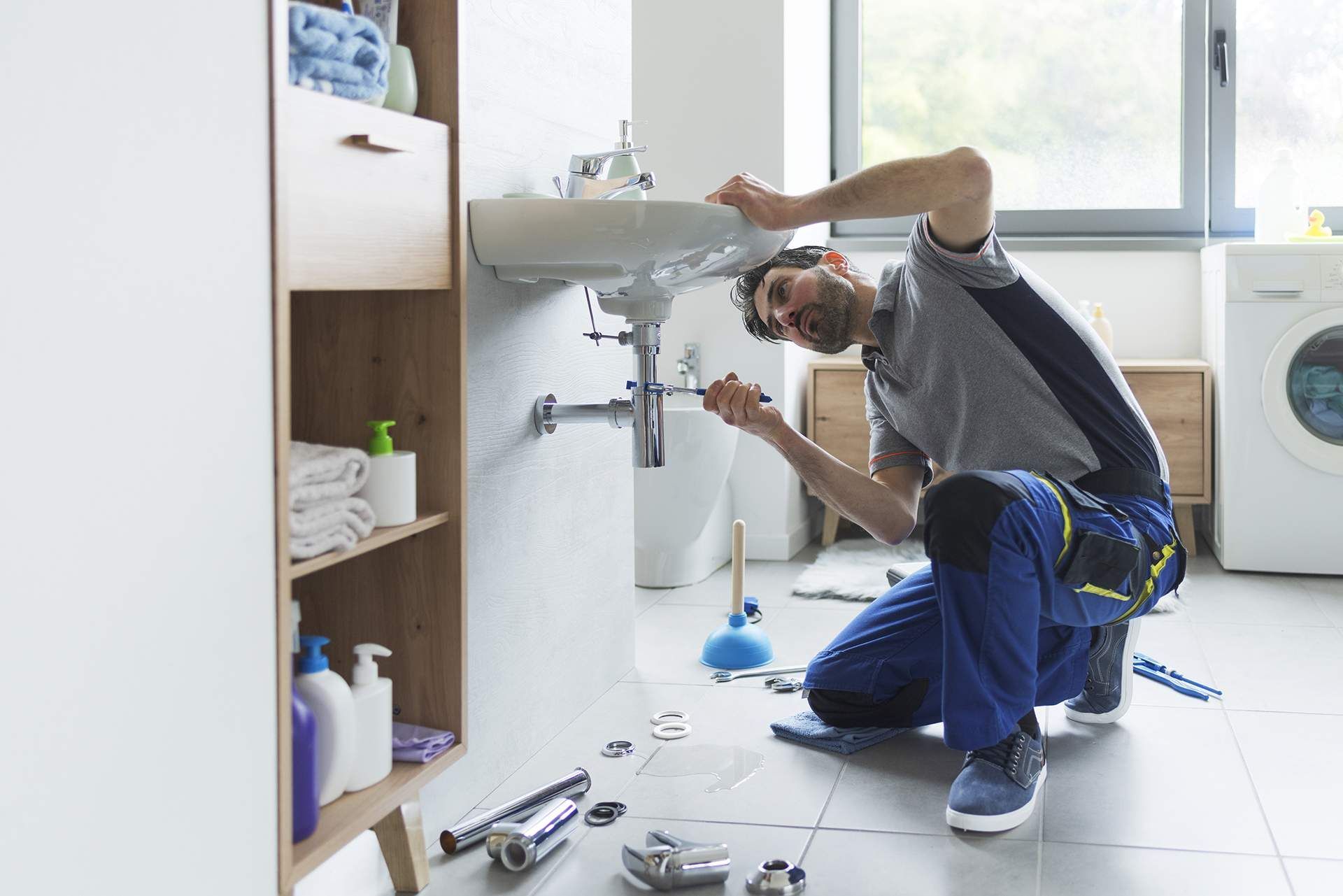 Plumber in blue overalls fixing sink in a bright bathroom, surrounded by tools, looking focused.
