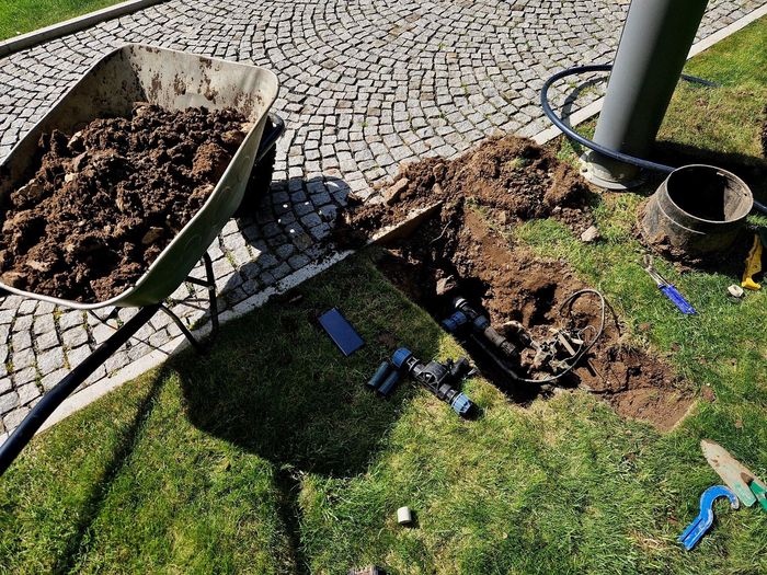 Wheelbarrow with dirt next to a trench being dug for irrigation pipes on a lawn.