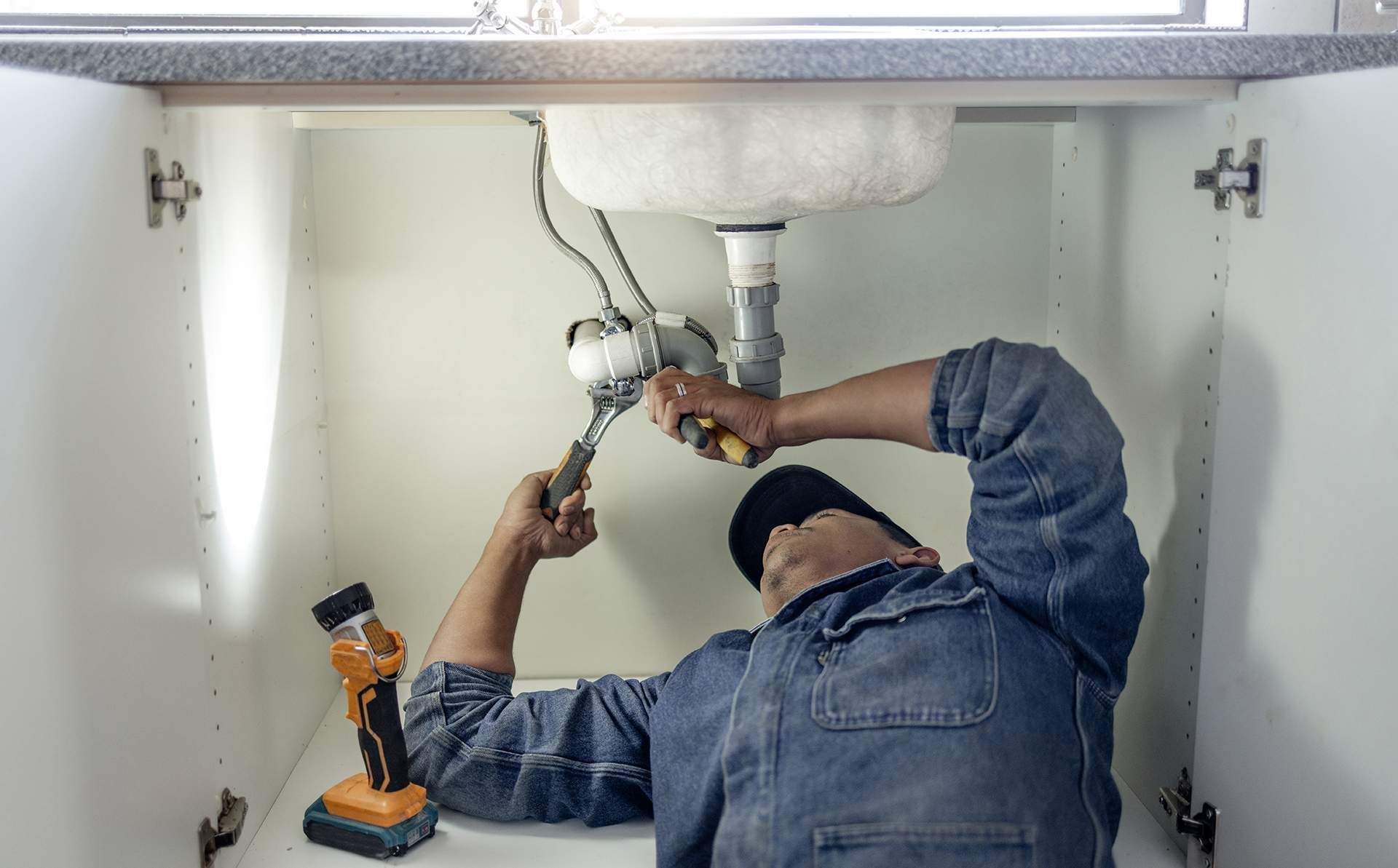 Plumber in blue denim shirt and black hat working under a sink with a wrench, in a white cabinet.